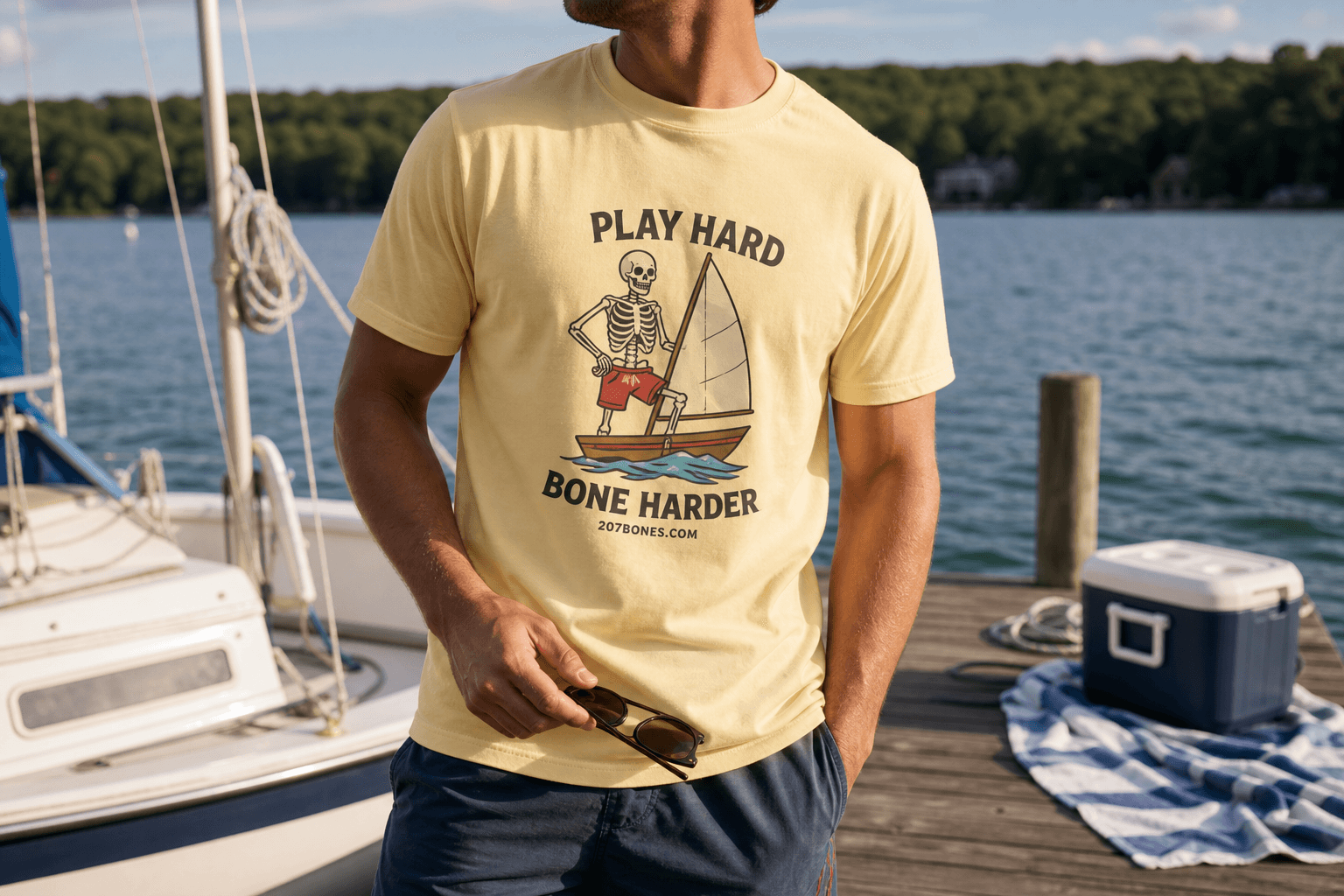 Adult wearing the pale yellow Play Hard Bone Harder sailing skeleton tee on a summer lake dock with their face mostly out of frame.
