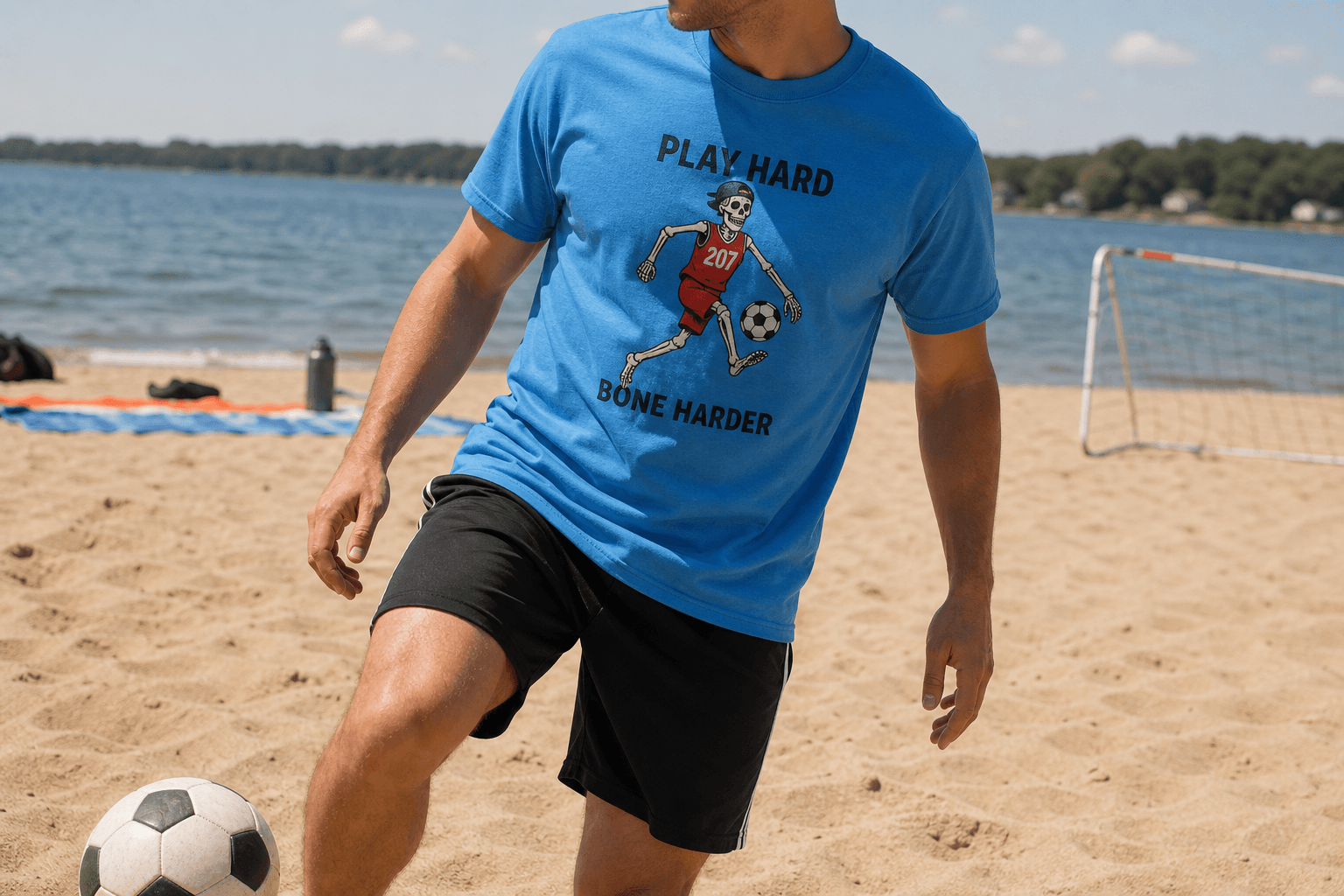 Adult wearing the blue Play Hard Bone Harder soccer skeleton tee during a beach soccer hangout with their face cropped out.