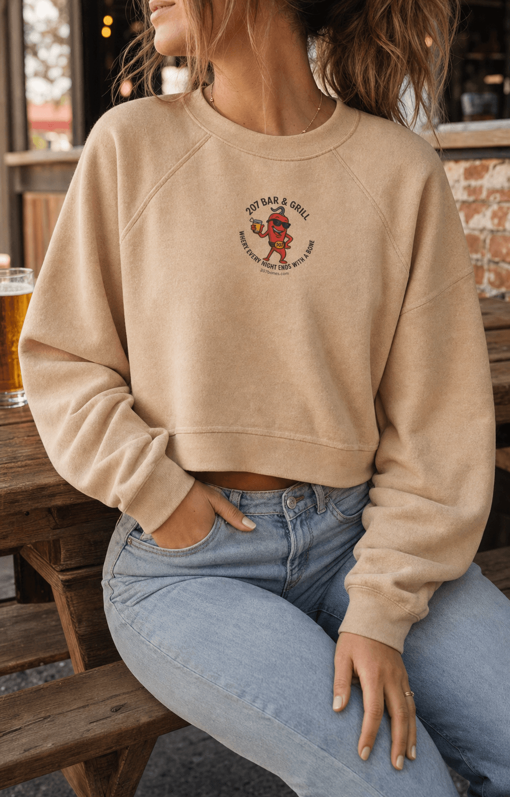 Adult woman wearing a tan cropped 207 Bar and Grill fleece pullover at a casual patio table.